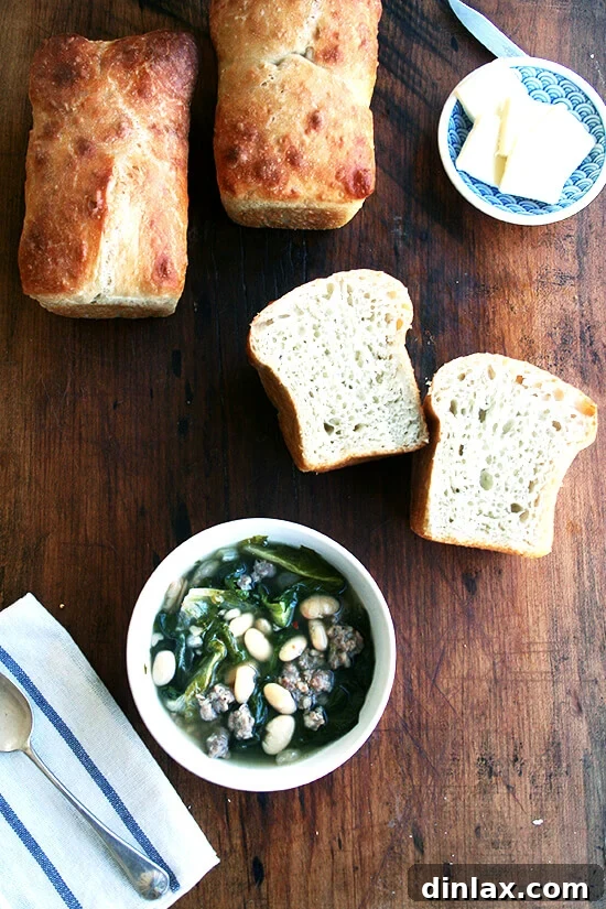 Perfectly paired: savory white bean soup with fresh, homemade peasant bread. A bowl of white bean, sausage, and escarole soup, served alongside slices of homemade peasant bread on a wooden board.