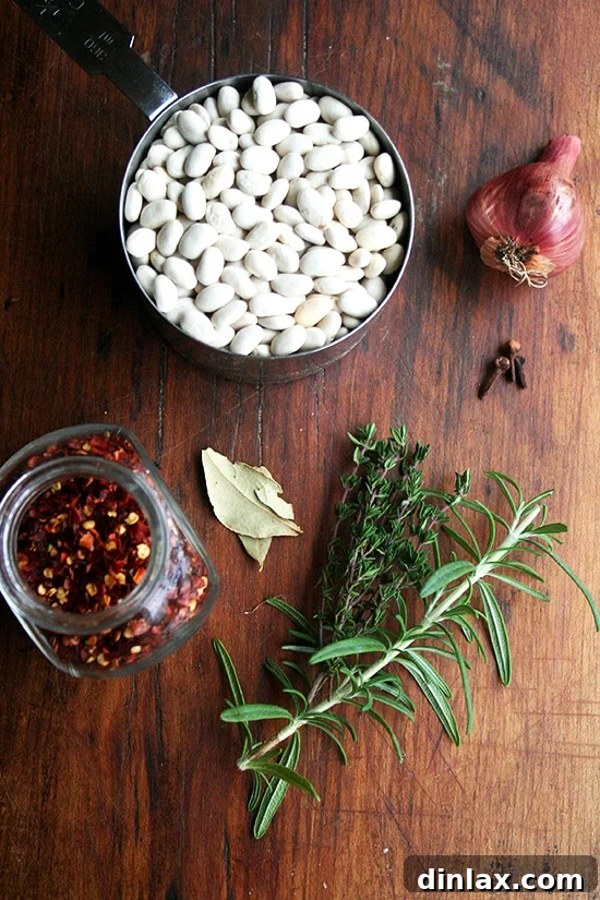 Hearty Italian Sausage, Escarole, and White Bean Soup 2 An assortment of dried white beans, alongside various dried herbs like rosemary and thyme, arranged on a rustic wooden board.
