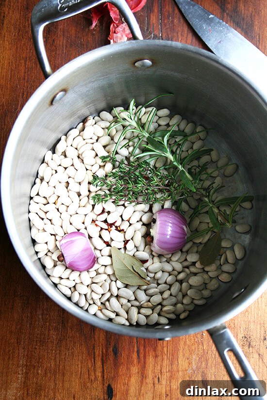 Hearty Italian Sausage, Escarole, and White Bean Soup 3 A large pot filled with dried white beans, aromatic herbs including a bay leaf, and a halved shallot, ready for cooking.