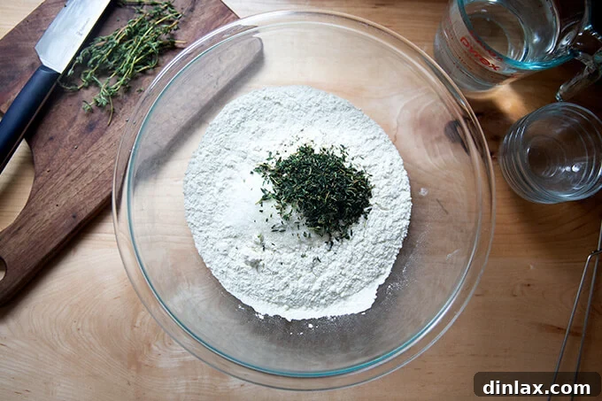 A bowl containing a mix of flour, salt, sugar, yeast, and fresh thyme leaves, ready to be combined.