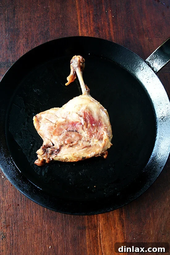 A duck leg confit sizzling skin-side down in a hot cast iron skillet, beginning to render fat and crisp up.