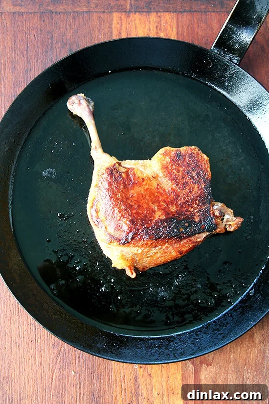 A beautifully seared duck leg confit with perfectly golden, crispy skin in a cast iron skillet, nearly ready to be served.