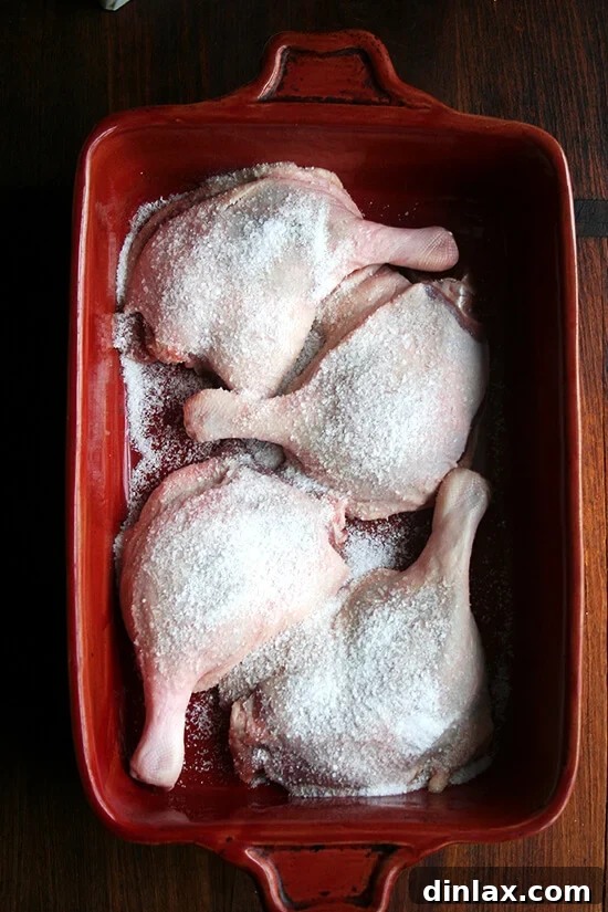 Four duck legs in a red baking dish, evenly sprinkled with kosher salt, ready for the next step.