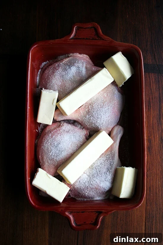 Four duck legs in a red baking dish, seasoned with salt and fully covered with slices of unsalted butter, ready for slow cooking.