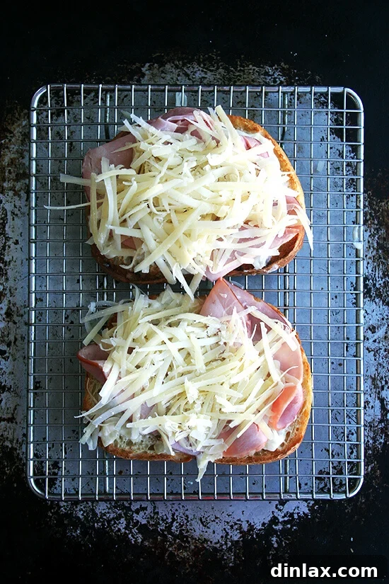 Croque monsieur in the making: Two slices of toast topped with béchamel, ham, and cheese on a rack set on a sheet pan.
