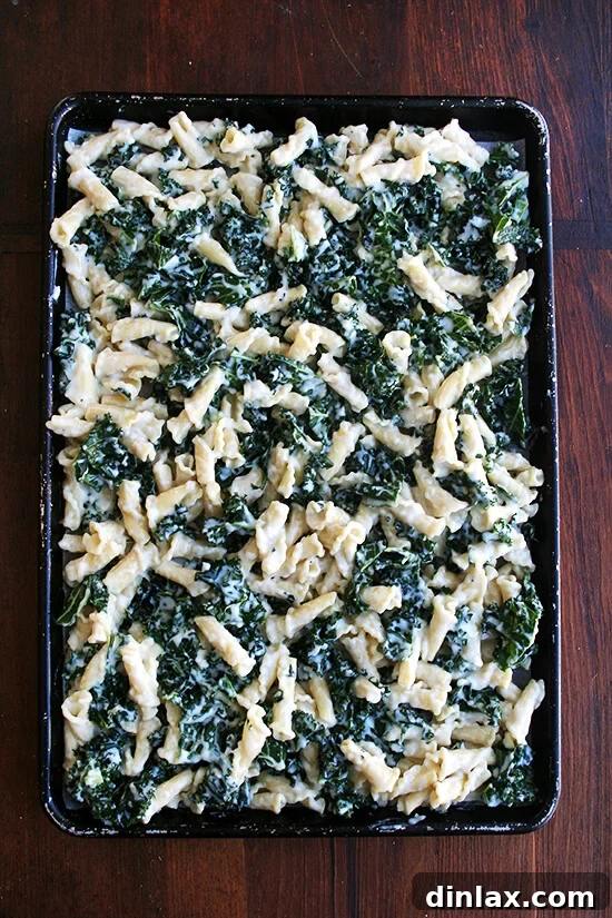 An overhead shot of unbaked pasta gratin with kale spread evenly on a parchment-lined sheet pan, topped with cheese cubes.