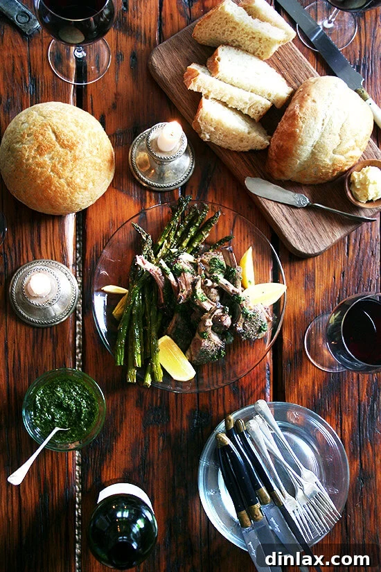 An overhead shot of a beautifully arranged Easter dinner table featuring homemade bread, a bowl of fresh mint sauce, perfectly broiled lamb chops, and a glass of red wine.