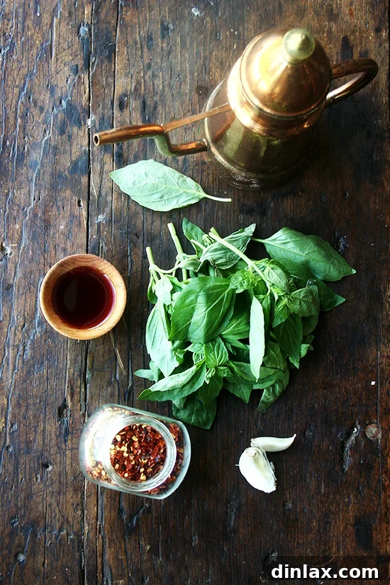 A rustic wooden board showcasing the key ingredients for the herb sauce: a bottle of olive oil, a small bowl of red wine vinegar, a pile of fresh green herbs (basil or parsley), minced garlic, and a pinch of red pepper flakes.