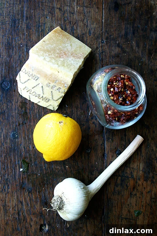 Tangy Swiss Chard Salad with Parmesan and Crunchy Breadcrumbs 4 Essential ingredients for the salad: wedges of Parmesan cheese, vibrant lemon, whole garlic cloves, and a small pile of red pepper flakes on a cutting board.