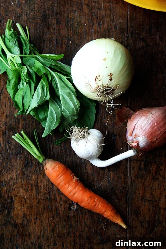 A vibrant assortment of fresh basil, onions, garlic, shallots, and carrots, neatly arranged on a table, ready for preparation.