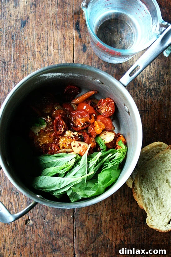 A saucepan on a stovetop, filled with the vibrant purée of roasted vegetables, ready to be blended. Adjacent are a measuring cup of water and slices of stale bread, crucial for the soup's unique texture.
