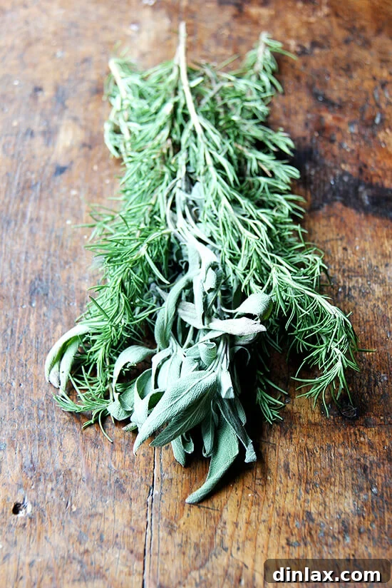 A rustic wooden board holds a few sprigs of freshly picked rosemary and sage, their vibrant green leaves promising aromatic flavor.