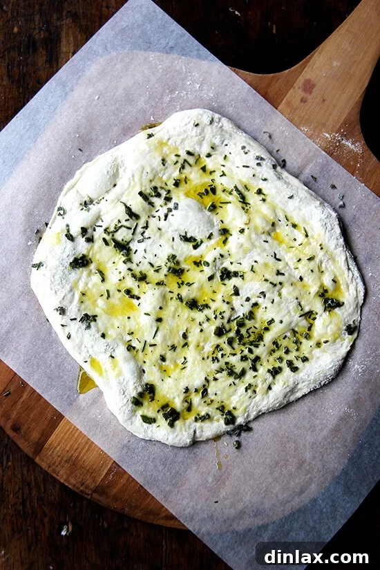 An unbaked flatbread, beautifully adorned with olive oil, sage, rosemary, and sea salt, rests on parchment paper on a baking peel, ready for the oven.