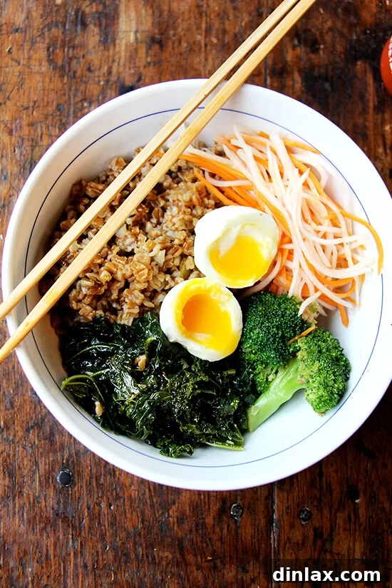 A vibrant leftover grain bowl featuring fluffy wheat berries, sautéed kale, steamed broccoli, bright quick-pickled carrots and daikon, drizzled with glossy teriyaki sauce, and topped with a perfectly soft-boiled egg with a runny yolk.