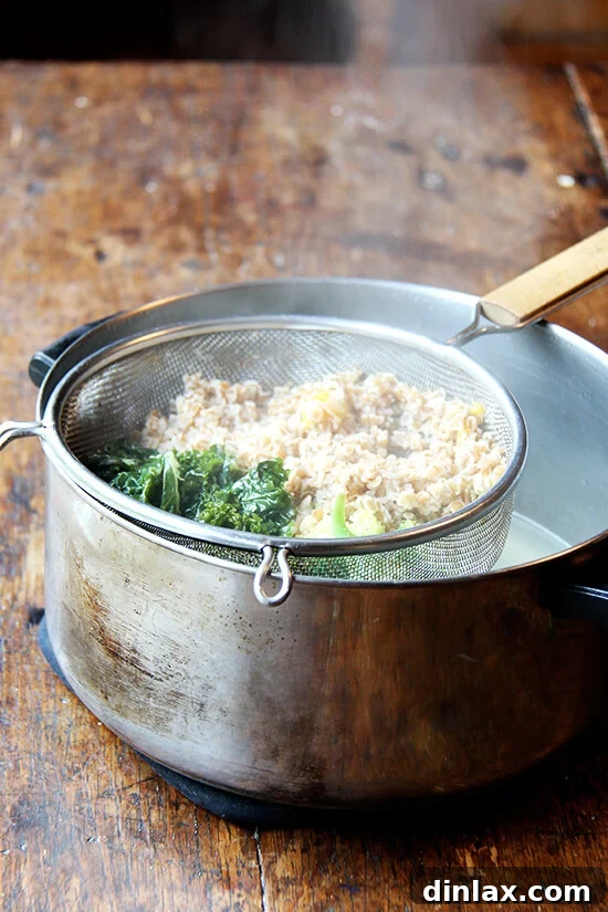 A large colander filled with cooked wheat berries and green vegetables, tightly covered with aluminum foil, positioned over a pot of simmering water for gentle and effective stovetop reheating.