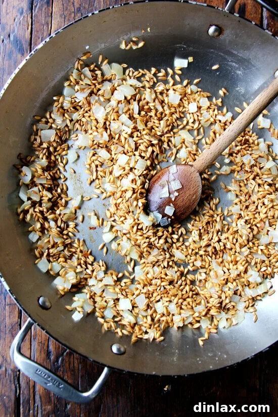 Hearty Farro Risotto with Autumn Squash and Kale 3 A wide sauté pan on a stovetop containing sautéed diced onion and pearled farro, starting to become translucent and fragrant, ready for the next step of the recipe.