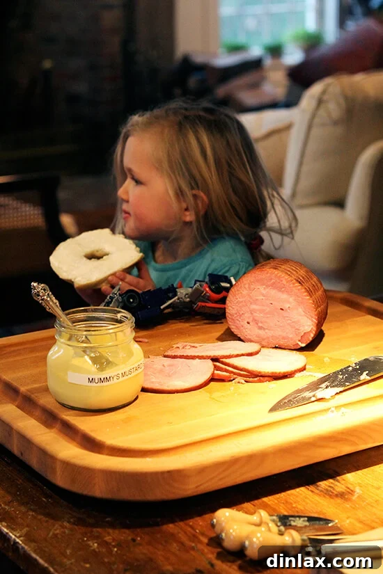 Ella enjoys a bagel, seated next to a serving board laden with delicious ham and a tangy mustard sauce.