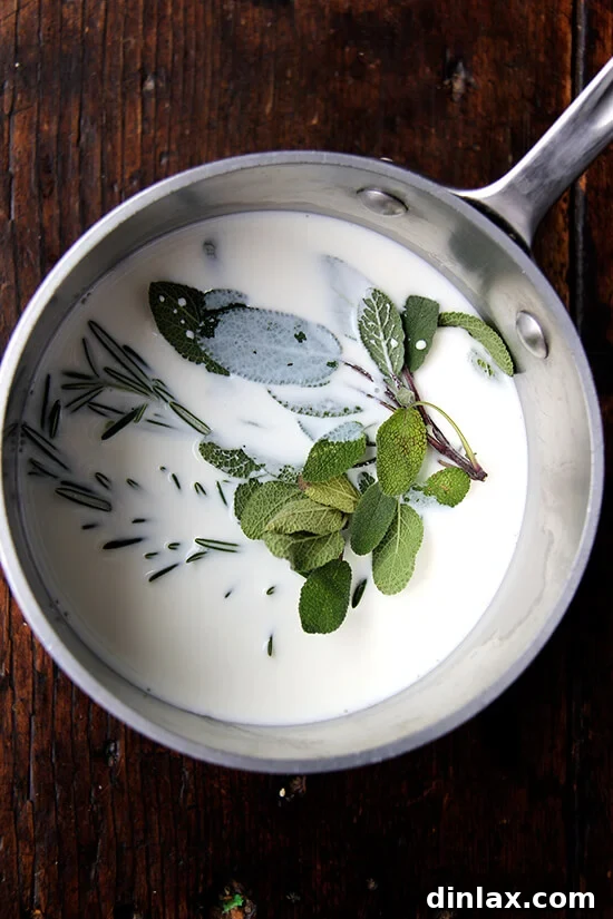 A saucepan filled with milk, fresh sage, and rosemary sprigs simmering on a stovetop.
