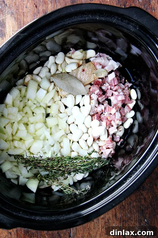 A slow cooker pot filled with a vibrant mixture of dried white beans, chopped onions, bay leaf, thyme sprigs, and a Parmesan rind, ready for the liquid ingredients.