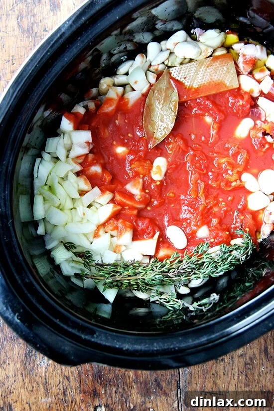 A slow cooker pot brimming with dried white beans, aromatic onions, fresh thyme, a bay leaf, and the deep red hues of crushed tomatoes, all combined for cooking.