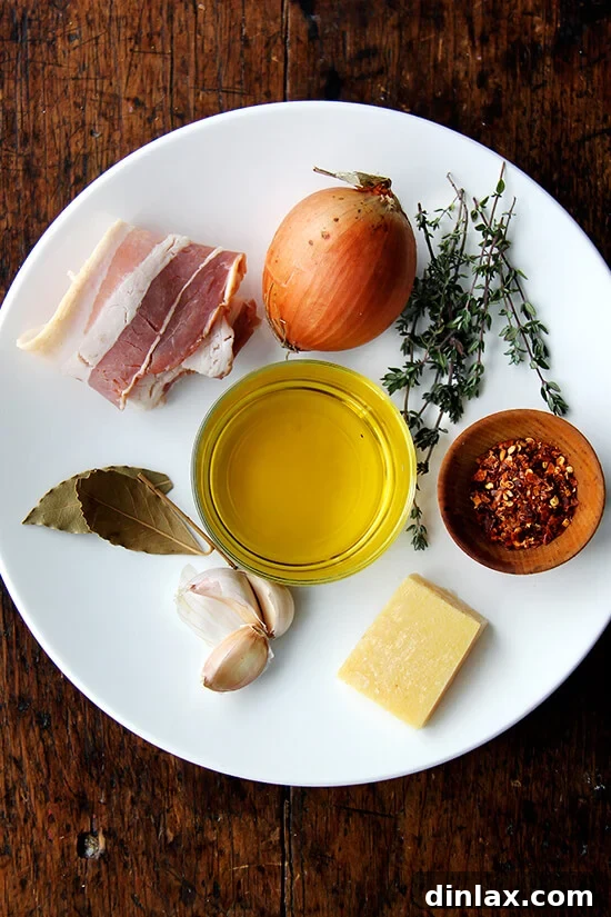A selection of ingredients for a flavorful bean broth including onions, garlic, bay leaves, thyme, olive oil, and bacon.
