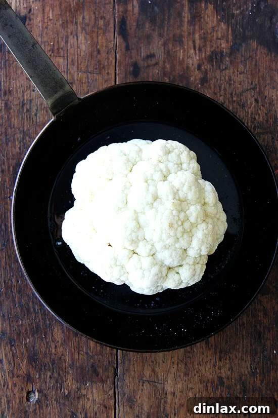 Whole cauliflower drizzled with oil and seasoning, placed in a sauté pan before roasting.