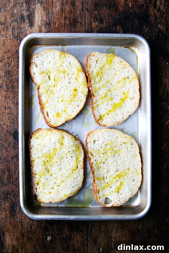 Briny Egg Salad Crostini 8 Slices of bread arranged on a baking sheet, drizzled with olive oil and ready for toasting in the oven.