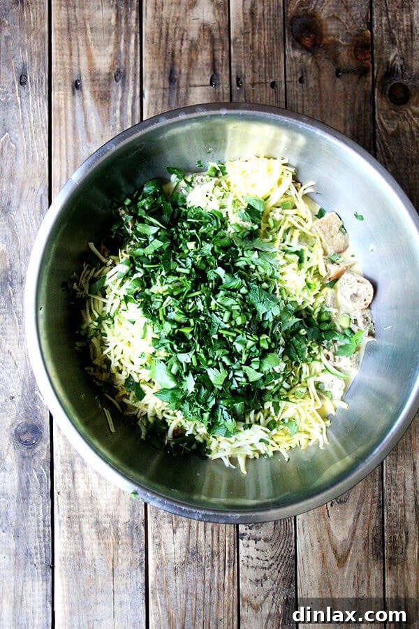 All the strata ingredients combined in a bowl: shredded vegetables, cheese, cubed bread, eggs, milk, and freshly chopped herbs like parsley and garlic scapes.