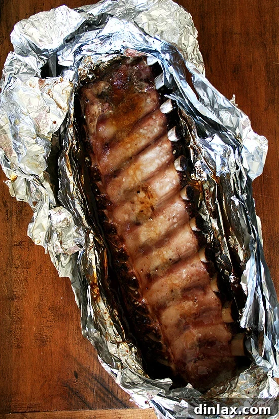 A full rack of perfectly oven-cooked baby back ribs, still sealed in aluminum foil, resting on a wooden cutting board just after being removed from the oven.