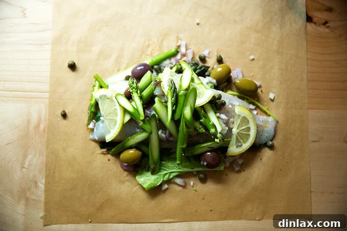 A sheet of parchment paper with a bed of Swiss chard, a branzino fillet, capers, and olives precisely arranged on it, awaiting the folding process.