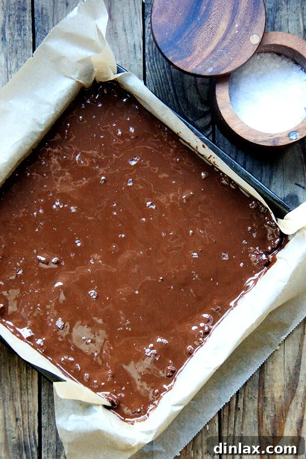 A delectable layer of rich chocolate ganache spread evenly over the crispy treats, elegantly sprinkled with flaky sea salt, chilling in the fridge to set perfectly.