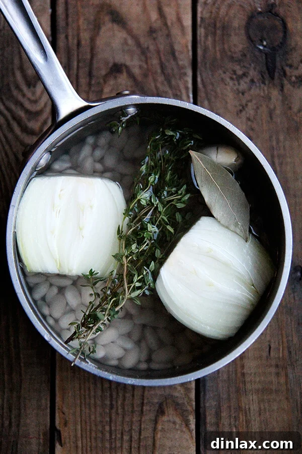 White beans covered in water with aromatics (onion, thyme, garlic, bay leaf) in a pot, demonstrating the start of the cooking process.
