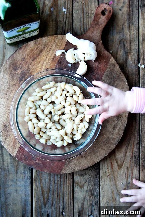 A selection of ingredients for stewy white beans laid out on a board: fresh garlic cloves, a bottle of olive oil, and a bowl of cooked white beans.