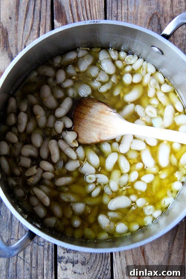 Cooked white beans being added to the saucepan with sautéed garlic and olive oil, ready to absorb the rich flavors.