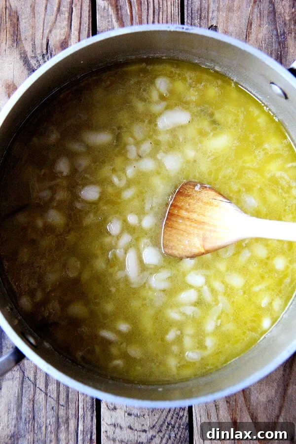 Brothy white beans simmering in a pot with a spoon, showing the creamy consistency developing as some beans break down.