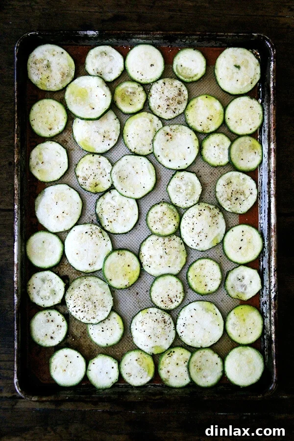 Sliced zucchini seasoned with salt, pepper, and olive oil, spread on a baking sheet.
