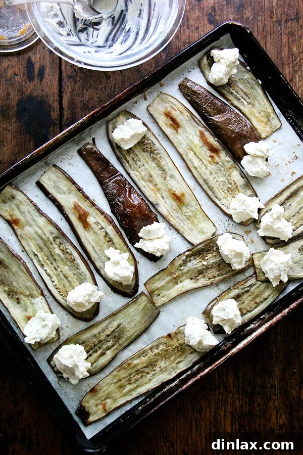 Roasted eggplant slices laid out on a sheet pan, each with a dollop of ricotta filling on one end, ready to be rolled into involtini.