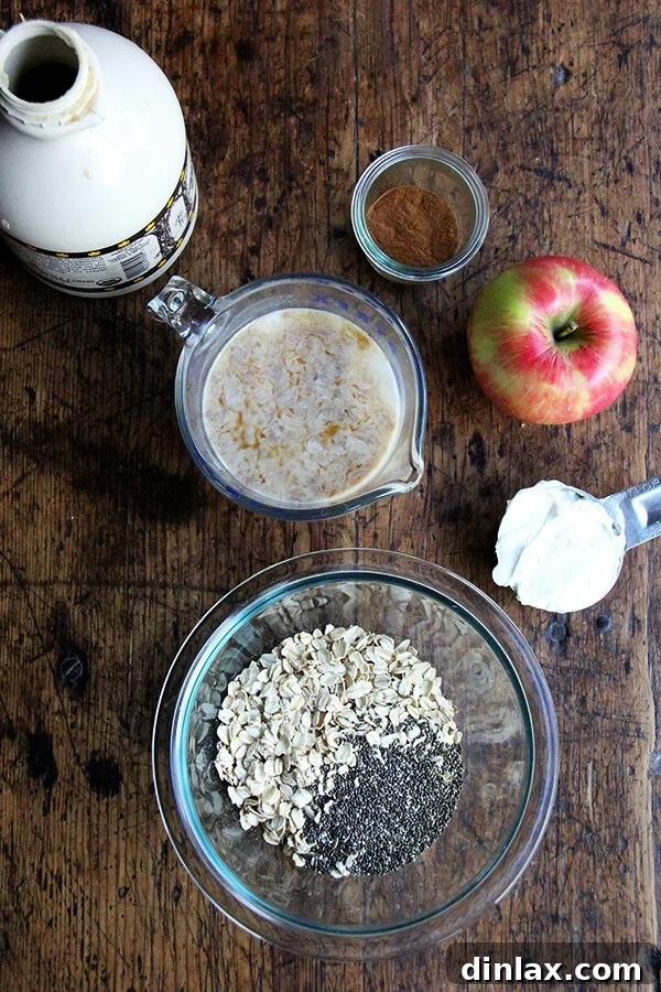 A collection of ingredients laid out for apple pie overnight oats, including rolled oats, almond milk, chia seeds, an apple, cinnamon, and maple syrup.
