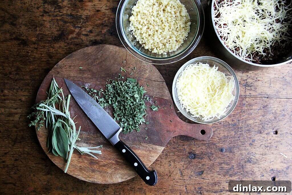 A cutting board laden with freshly chopped sage, rosemary, and thyme, alongside bowls of bread crumbs, shredded Gruyère and Parmesan cheese, and cooked quinoa, illustrating the preparation process.
