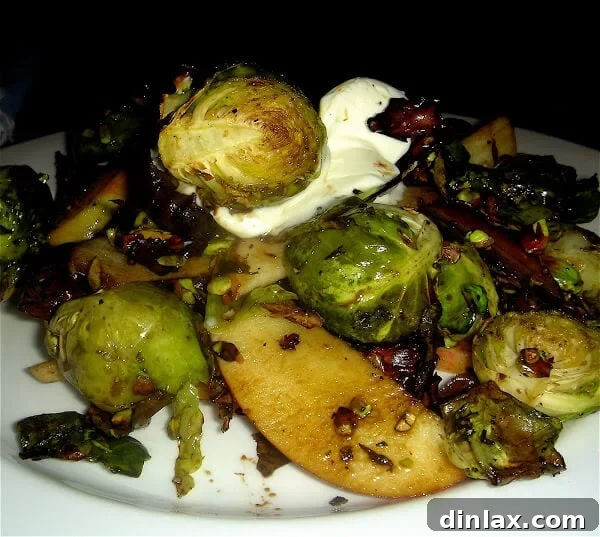 A serving of roasted Brussels sprouts, vibrant green with golden-brown edges, elegantly arranged with thin apple slices, dollops of crème fraîche, and a sprinkle of chopped pistachios.