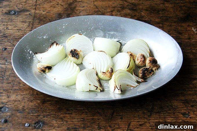 Yellow onions and garlic cloves broiling on a sheet pan, acquiring charred spots for smoky flavor.