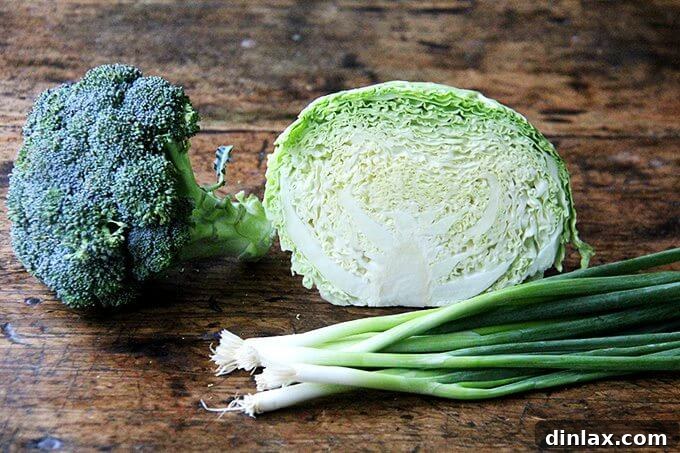 Fresh ingredients laid out on a cutting board: a head of vibrant green broccoli, a crisp white cabbage, and several bright green scallions.