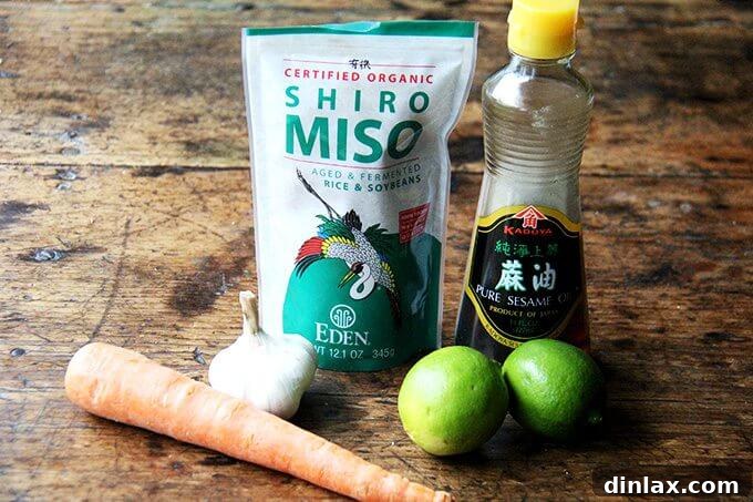 The key ingredients for the vibrant miso-carrot dressing are arranged on a rustic wooden table: fresh carrots, a container of white miso paste, garlic cloves, bright green limes, and a bottle of sesame oil.
