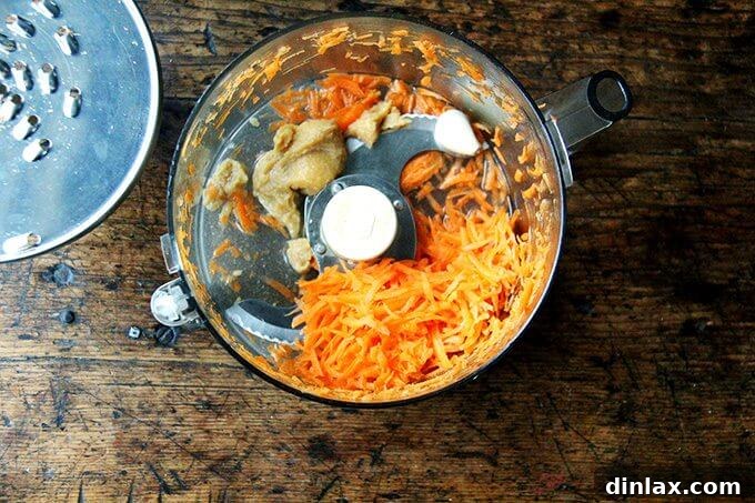 A food processor bowl containing freshly shredded carrots, creamy white miso paste, and garlic cloves, ready to be blended into a flavorful dressing.
