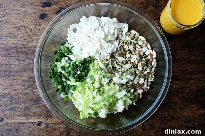 A large bowl containing all the fresh ingredients for the cabbage slaw: shredded cabbage, cauliflower, vibrant green scallions, toasted almonds, and bright green pepitas.