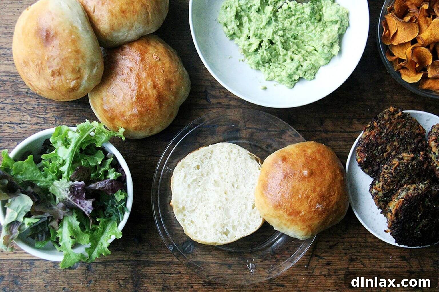 A beautifully arranged wooden board featuring fluffy homemade brioche buns, a bowl of creamy mashed avocado, and several cooked mushroom-quinoa burger patties, ready for assembly.