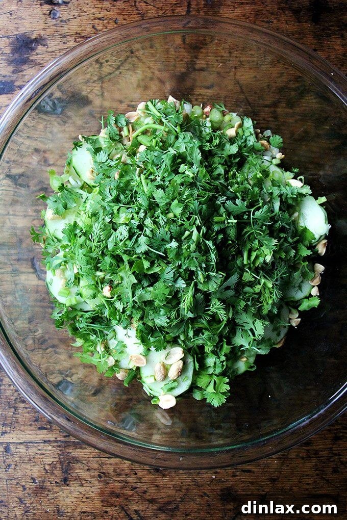 A large bowl filled with crisp cucumber slices, fresh cilantro, mint leaves, roasted peanuts, and vibrant lime wedges, awaiting to be tossed into a Vietnamese cucumber salad.