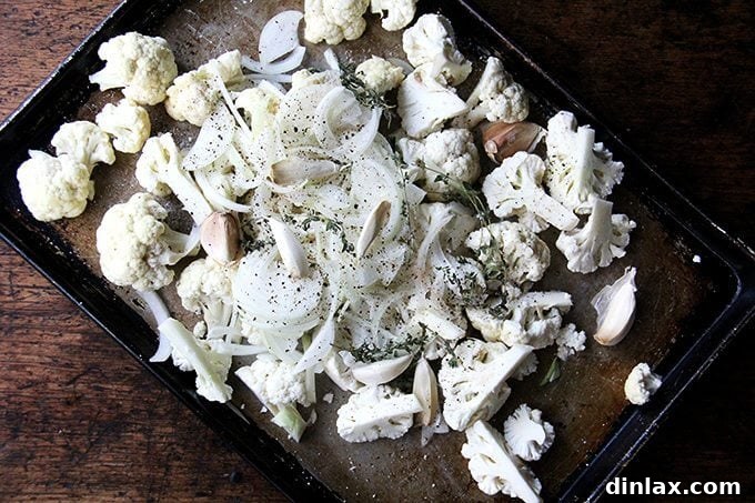 A sheet pan loaded with fresh cauliflower florets, sliced onions, unpeeled garlic cloves, and sprigs of thyme, seasoned and ready for roasting.