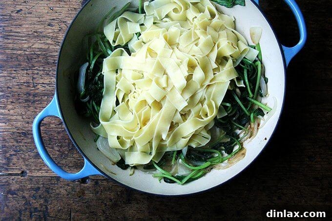 An overhead view of a skillet containing sautéed greens and perfectly cooked pasta, ready for the carbonara sauce.