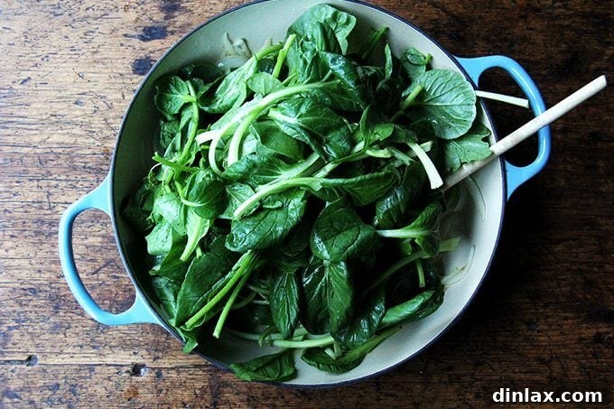 An overhead view of a skillet now filled with fresh, dark leafy greens added to the softened onions, ready to be wilted.